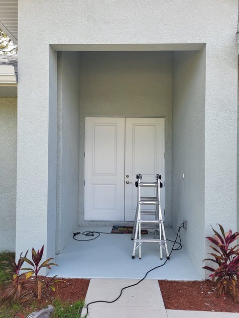 Garage entryway with white double doors, aluminum ladder leaning against wall, and potted plants on sides