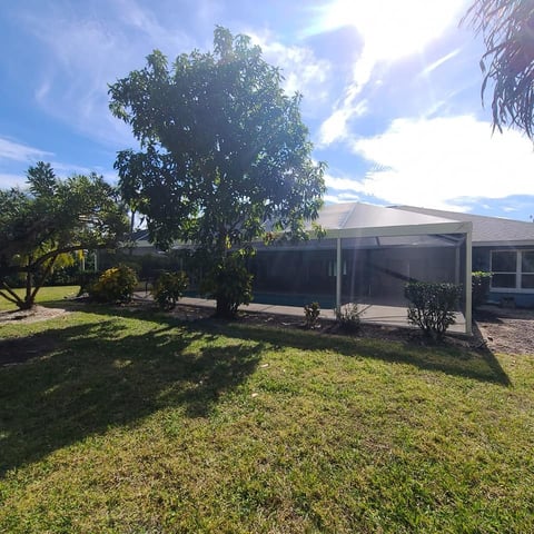 Single-story residential house with carport and shade structures surrounded by green lawn and mature trees on a sunny day