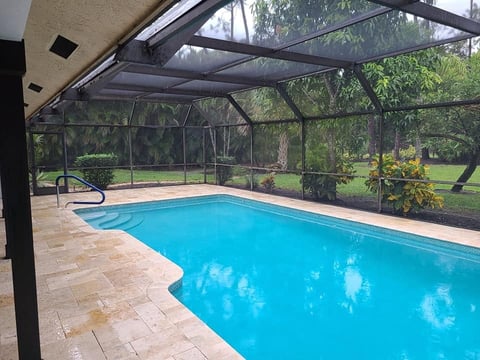 Screened-in backyard swimming pool with bright blue water, beige patio deck, and lush green lawn visible beyond the enclosure