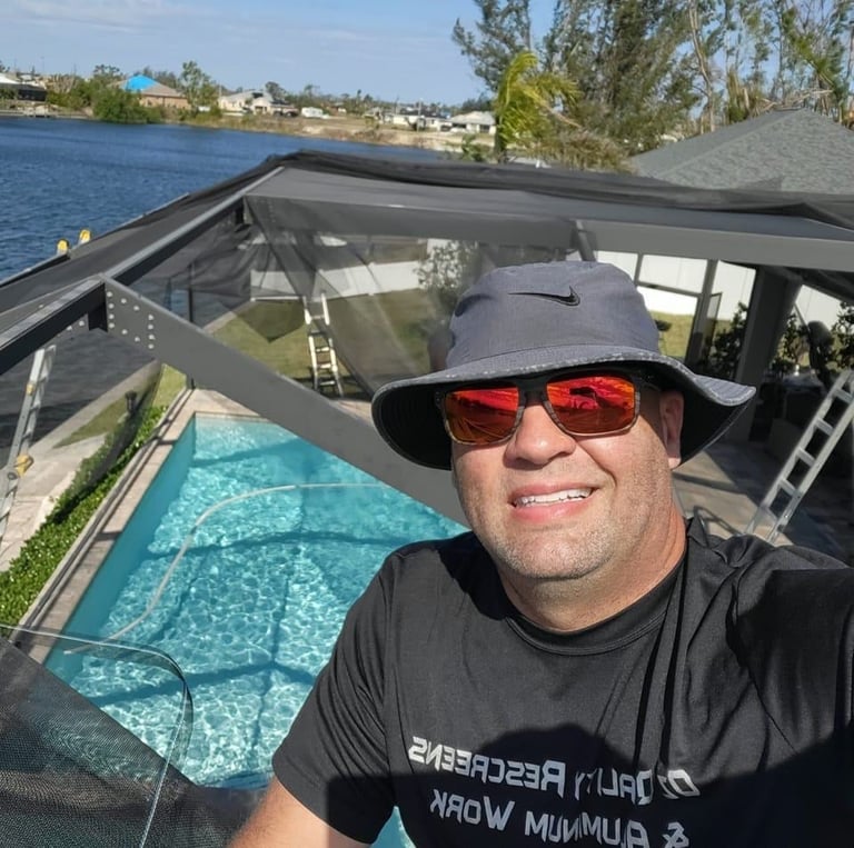 Man wearing sunglasses and black hat taking selfie near pool with waterfront and moored boat in background